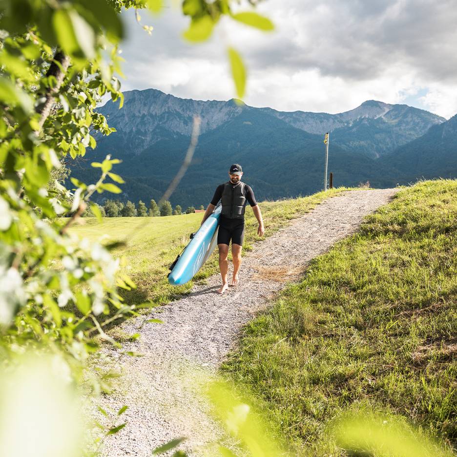 Muž v neoprénovom obleku a záchrannej veste nesie paddleboard v horskej krajine.