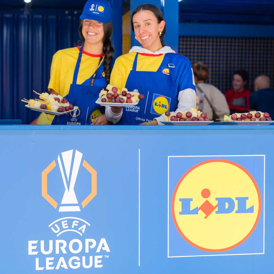 Dve usmievavé ženy v zásterách Lidl ponúkajú ovocie na špajdliach na podujatí UEFA Europa League.