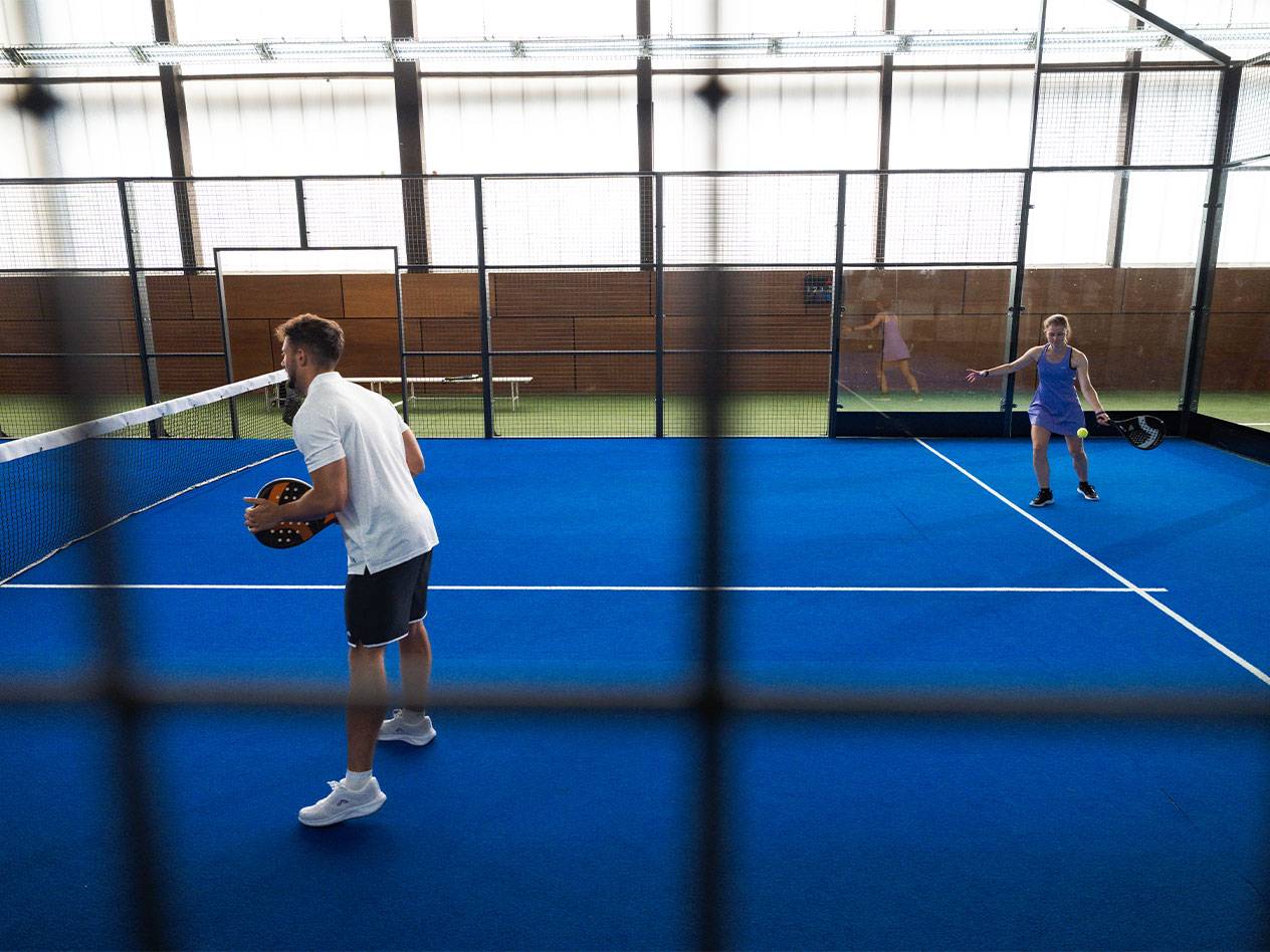 Muž a žena hrajúci padel na modrom kurte s raketami a loptičkou.