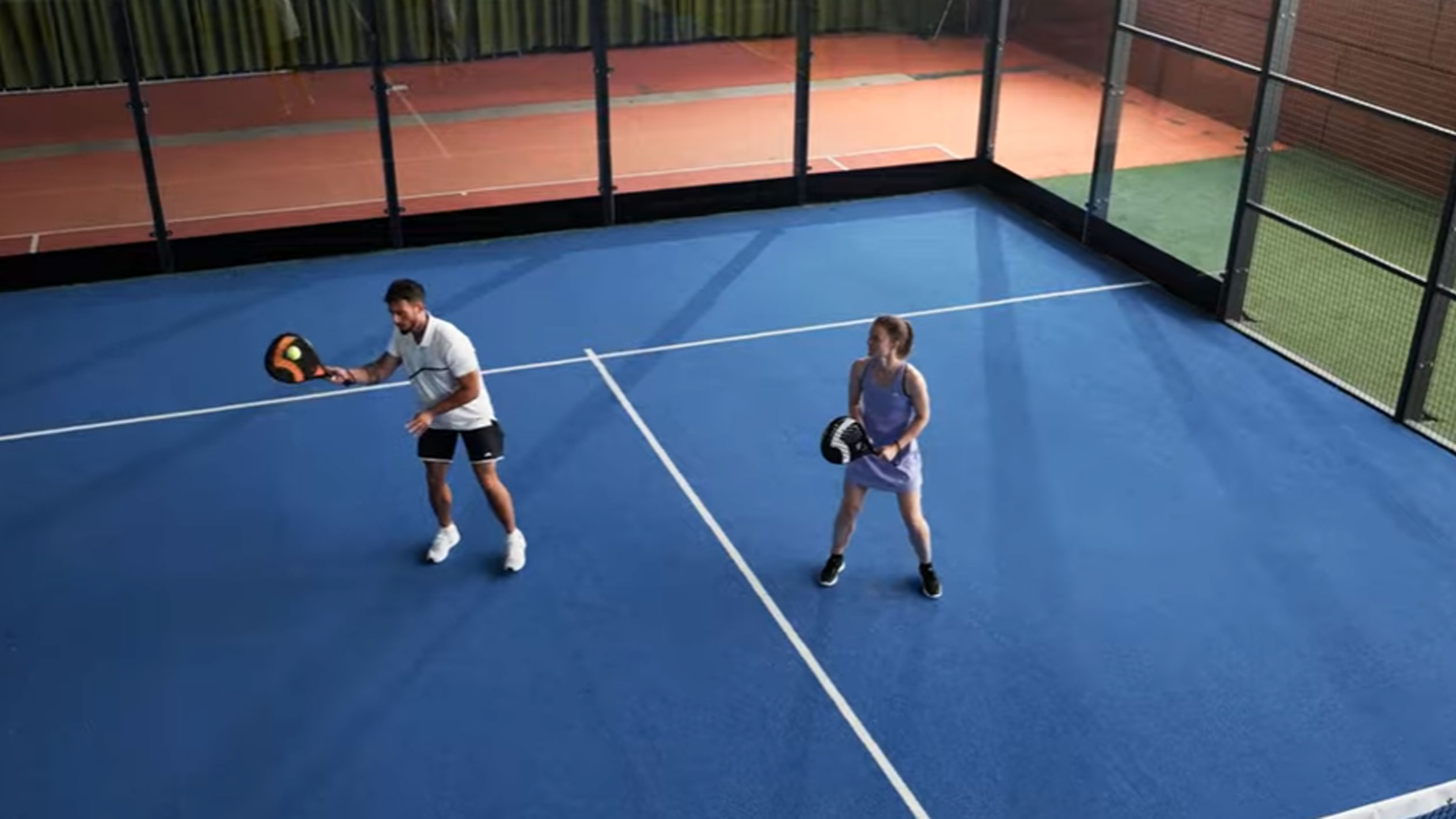 Muž a žena hrajú padel na modrom kurte s raketami a loptičkou