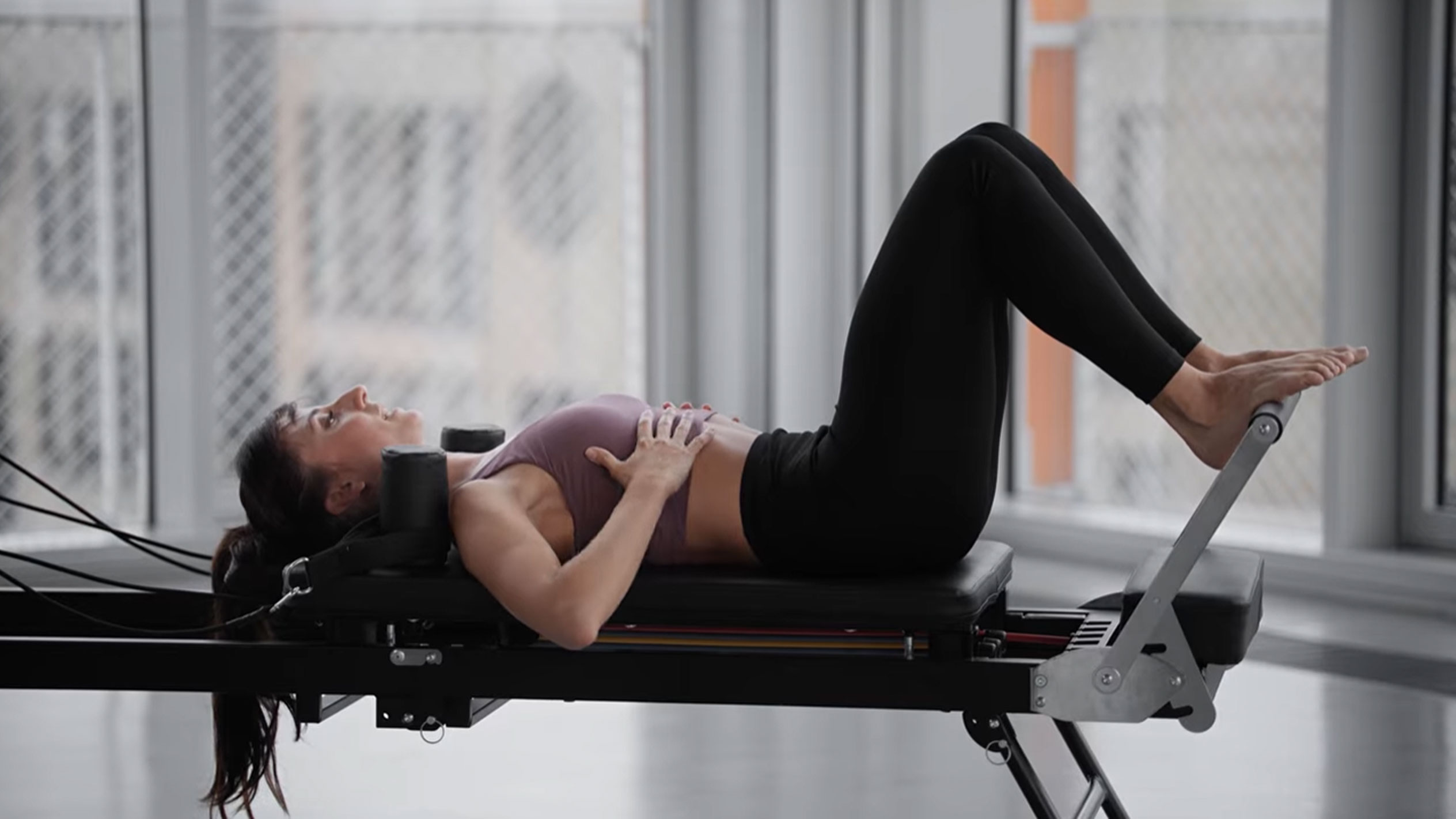 Žena cvičiaca pilates na reformeri, s rukami na hrudi.