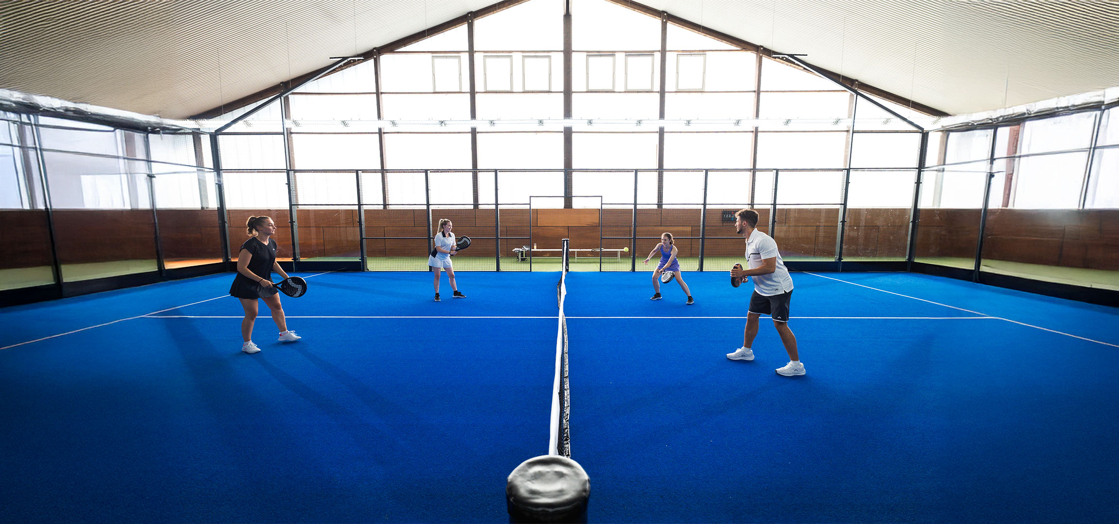 Štyria ľudia hrajú padel na modrom kurte, s raketami a športovým oblečením.