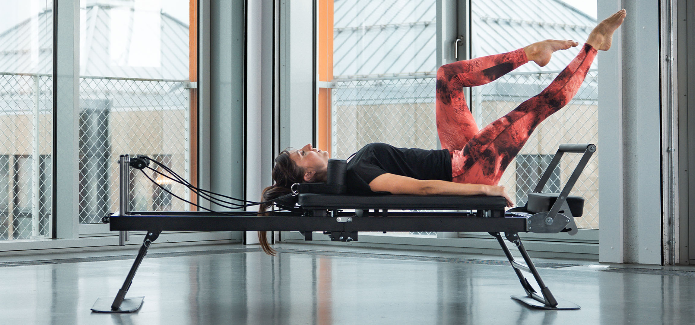 Žena cvičiaca Pilates na reformer stroji, s fitness doplnkami v blízkosti.