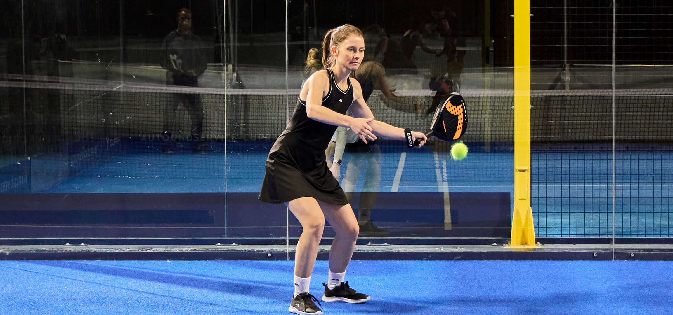 Žena hrajúca padel na modrom kurte, oblečená v čiernom športovom oblečení.