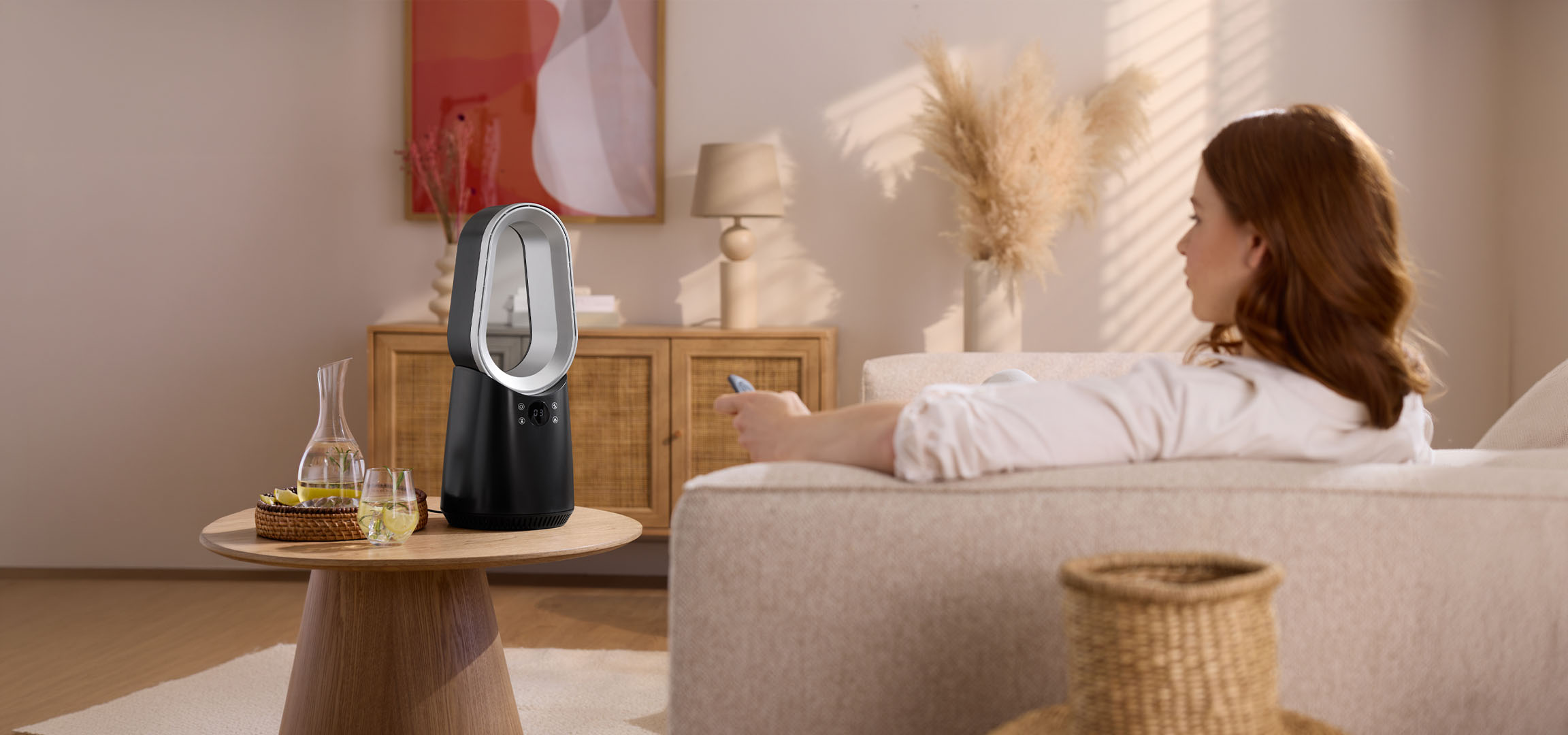 Žena na gauči s diaľkovým ovládačom, moderný ventilátor a nápoje na stole.