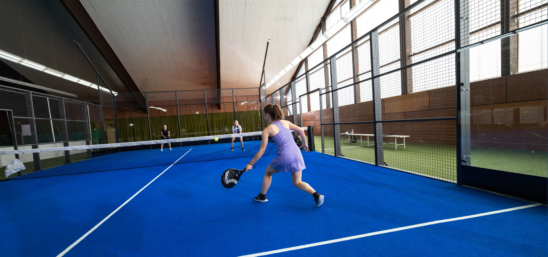 Žena hrajúca padel na modrom kurte, s ďalšími hráčmi v pozadí.