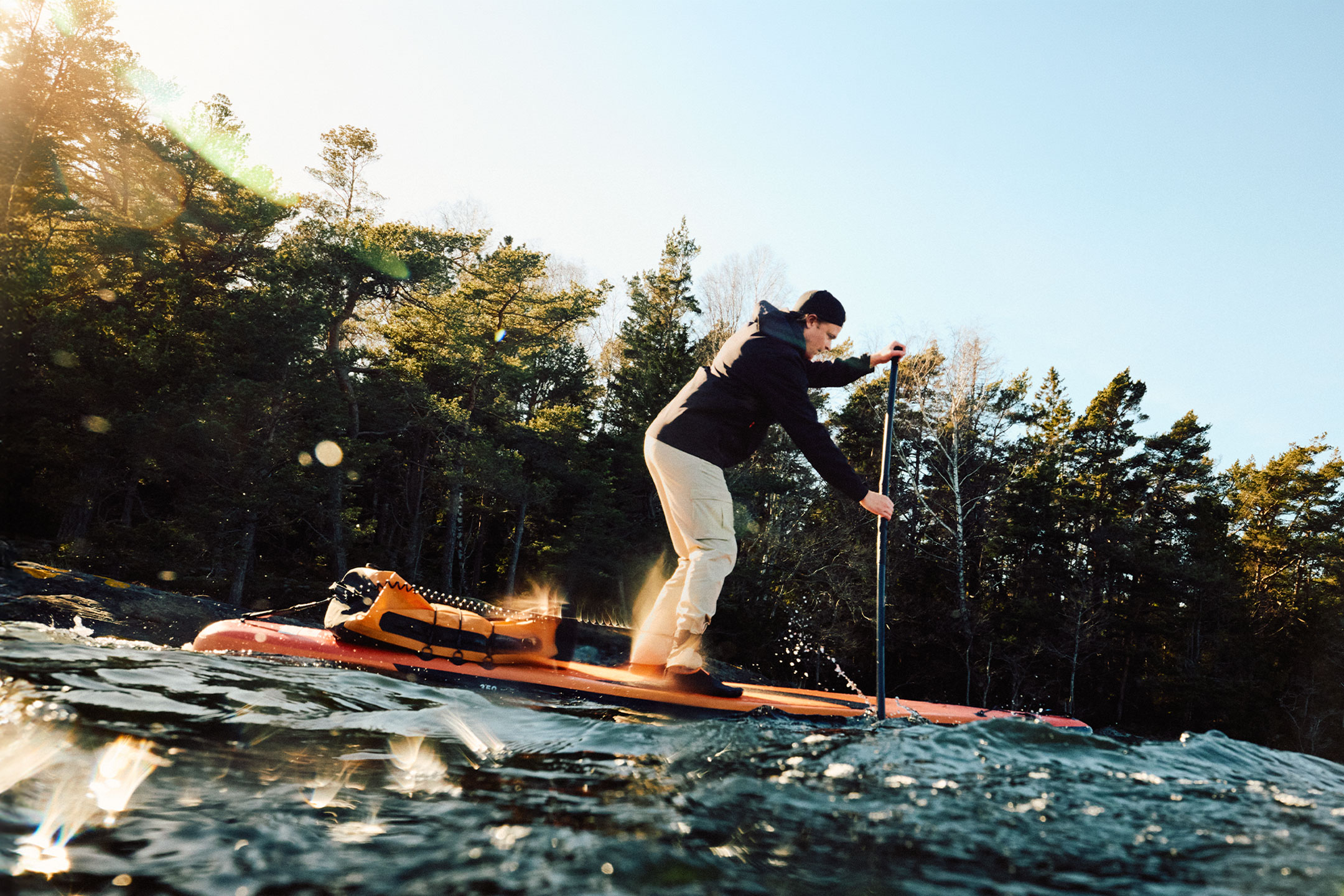 Muž na paddleboarde s oranžovou suchou taškou pri lese.