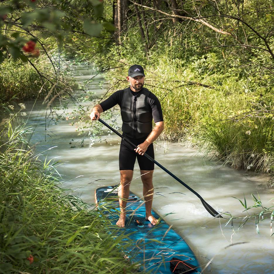 Muž v neoprénovom obleku a záchrannej veste stojí na paddleboarde v potoku.