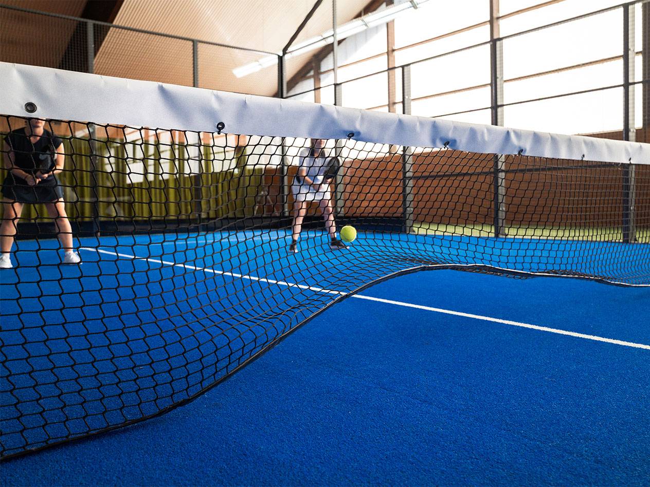 Dve ženy hrajú padel na modrom kurte, so sieťou v popredí.
