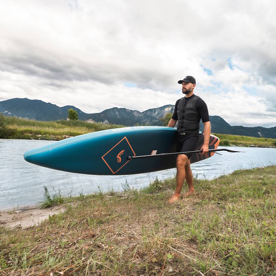 Muž v záchrannej veste a čiapke nesie paddleboard a pádlo pri rieke.
