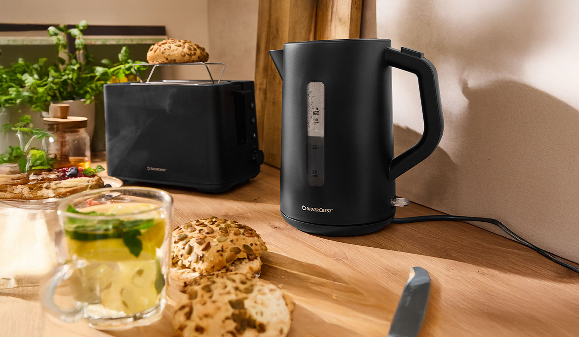 Čierna rýchlovarná kanvica a hriankovač SilverCrest na drevenom stole s raňajkami.