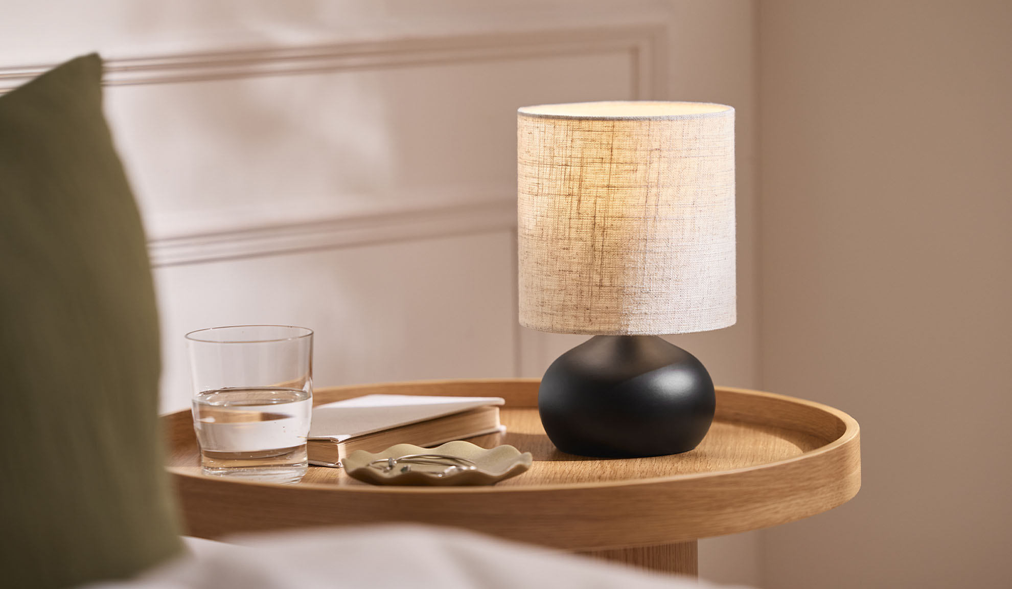 Nočná lampa, pohár, kniha a šperkovnica na drevenom podnose vedľa postele.