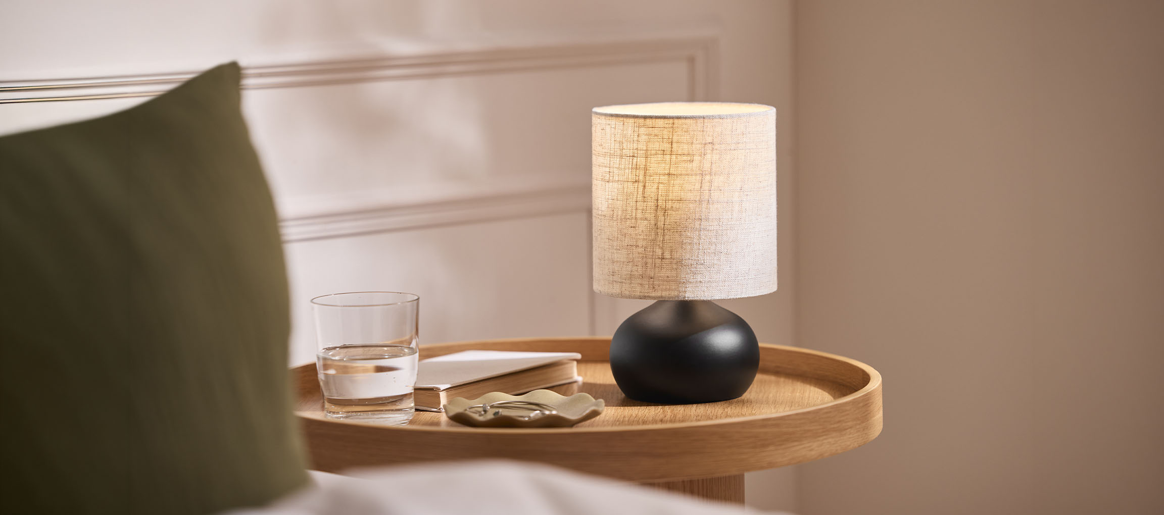 Nočná lampa s látkovým tienidlom, pohár vody a kniha na drevenom podnose.