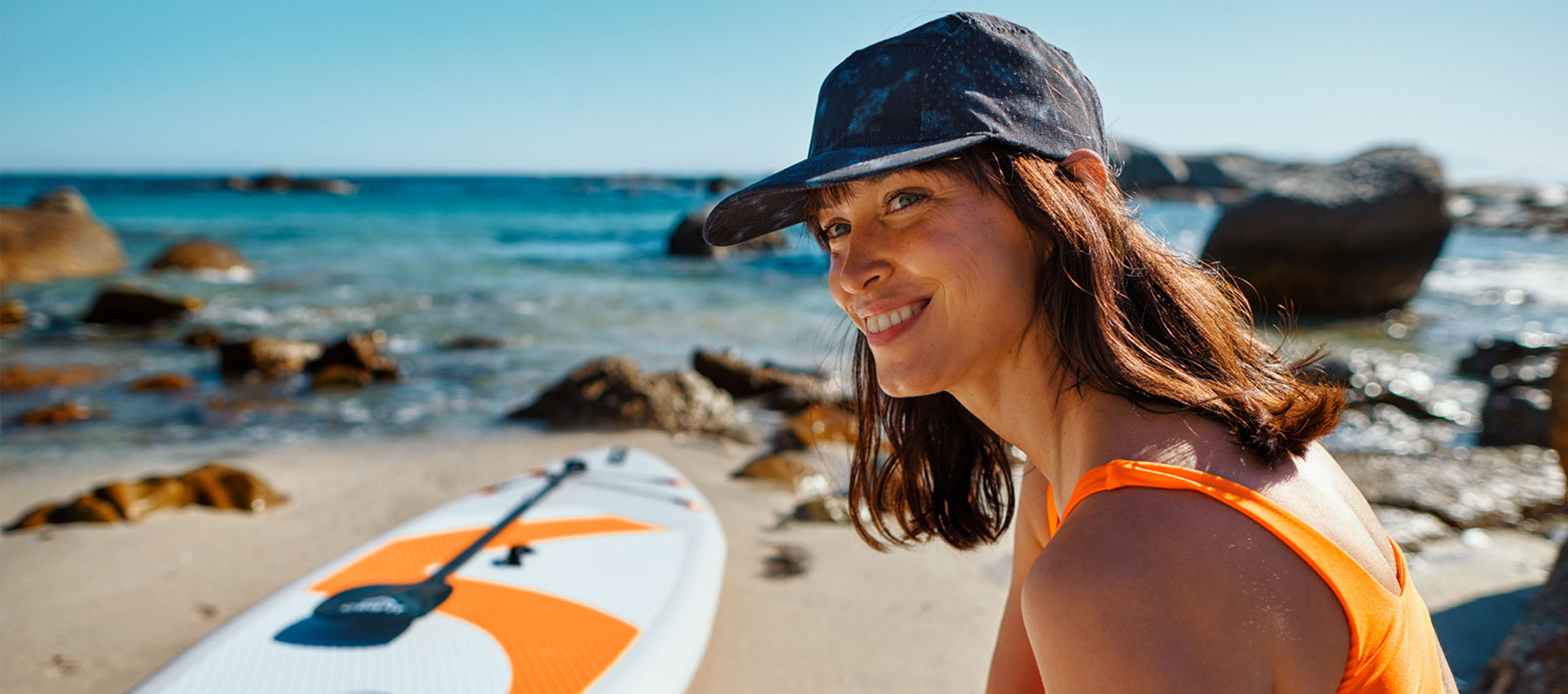 Usmiata žena v oranžových plavkách a baseballovej čiapke pri paddleboarde na pláži.