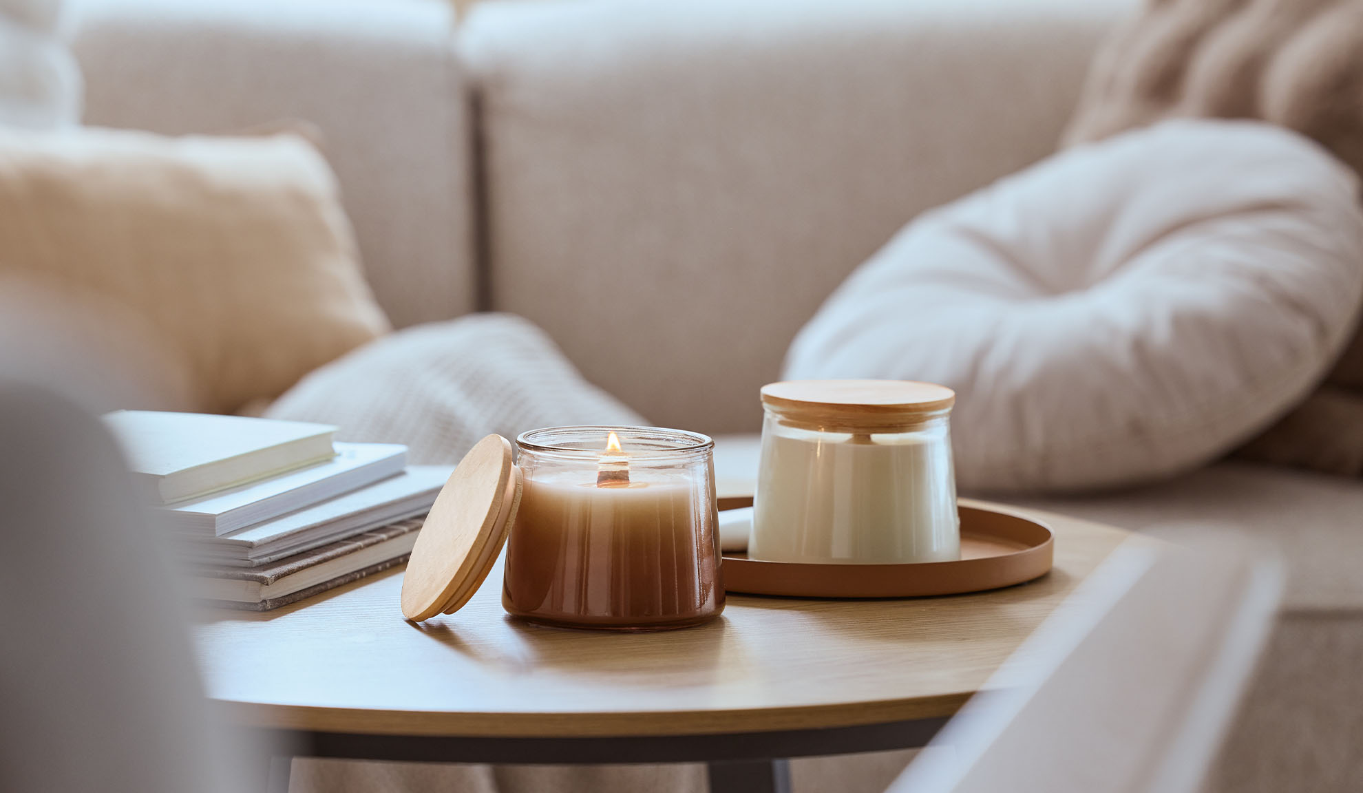 Dve vonné sviečky na drevenom stole, jedna zapálená, s útulnou pohovkou v pozadí.