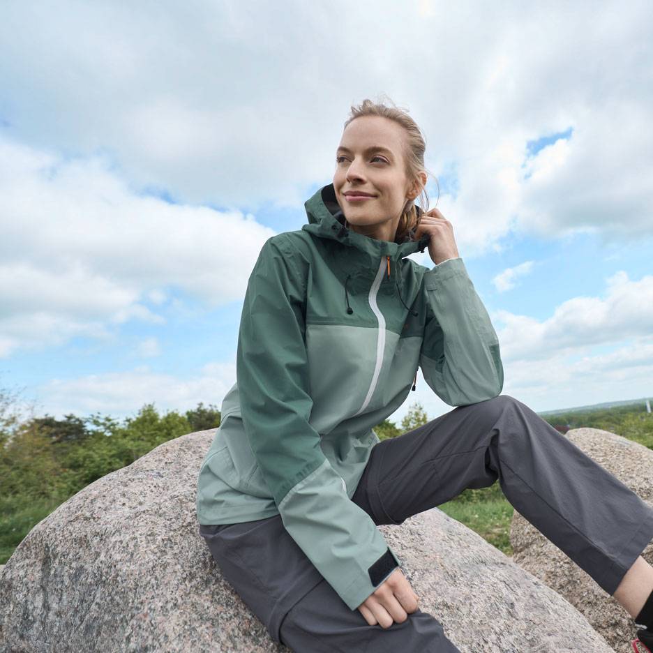 Žena v zelenej outdoorovej bunde a sivých nohaviciach sedí na skale a pozerá do diaľky.