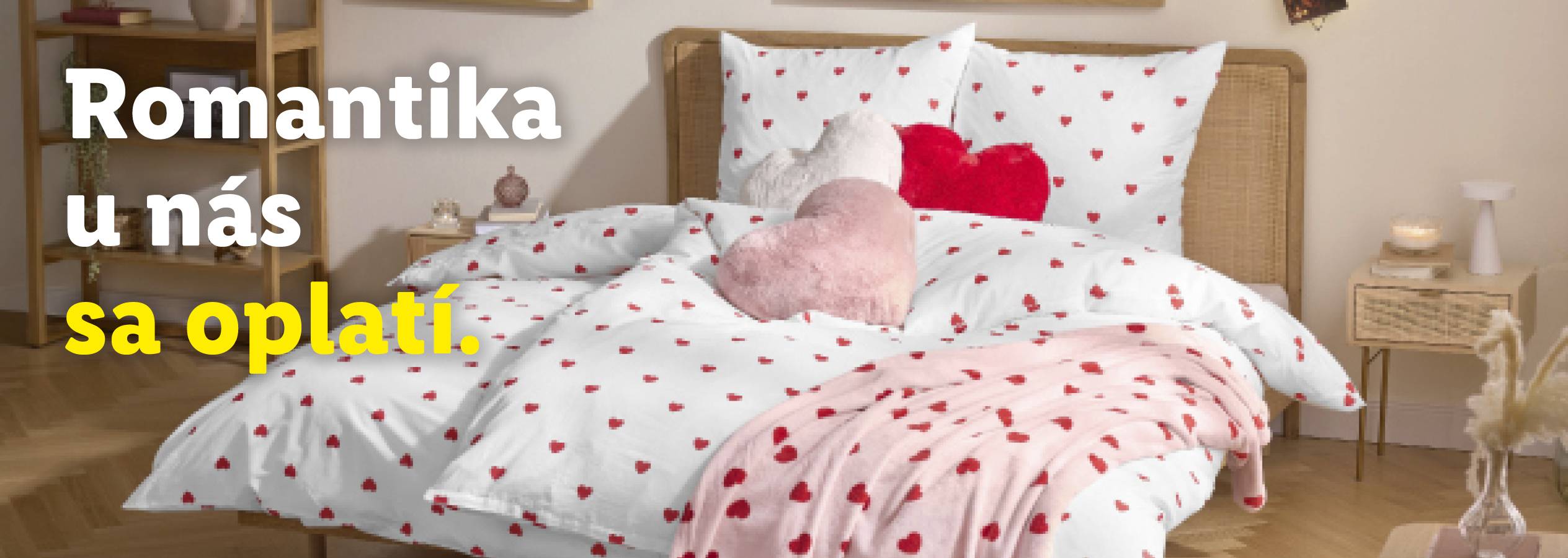 Posteľ s bielou posteľnou bielizňou s červenými srdiečkami a vankúšmi v tvare srdca. Text: Romantika u nás sa oplatí.