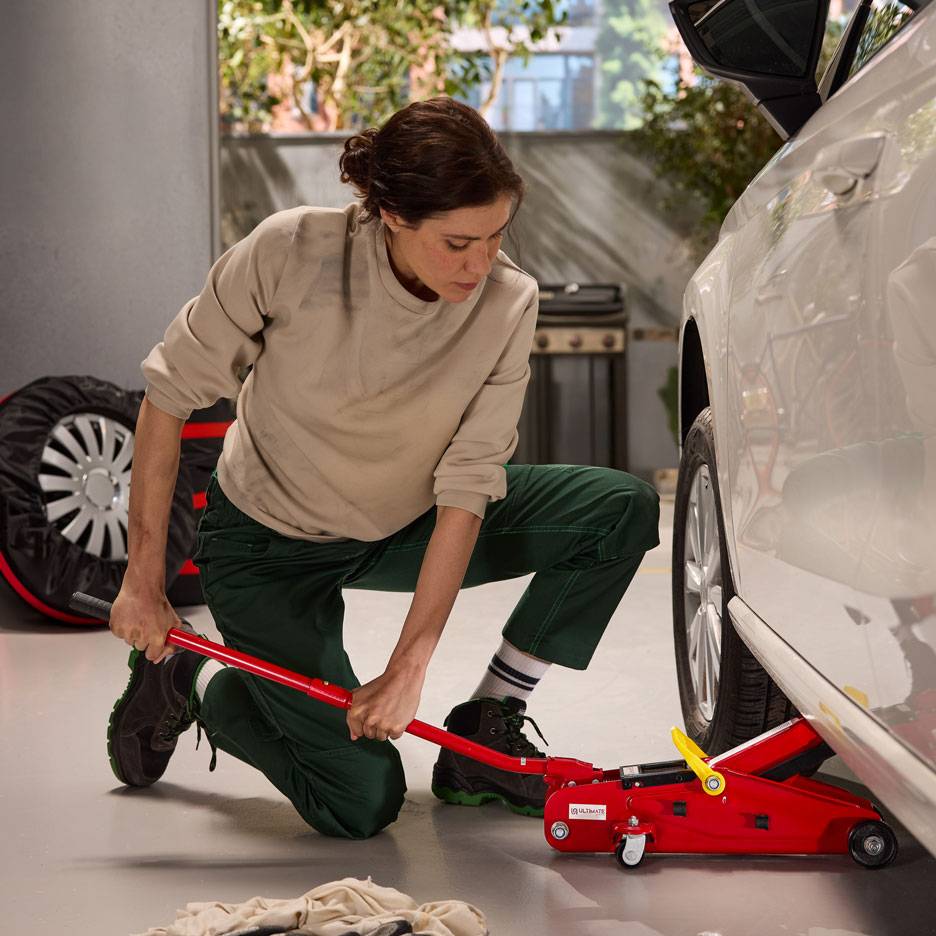 Žena používa červený hydraulický zdvihák na zdvíhanie bieleho auta, s rezervnou pneumatikou nablízku.