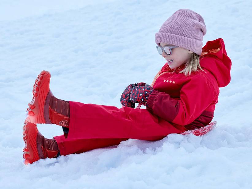 Dieťa v červenom overalí a červených čižmách sa sánkuje v snehu.
