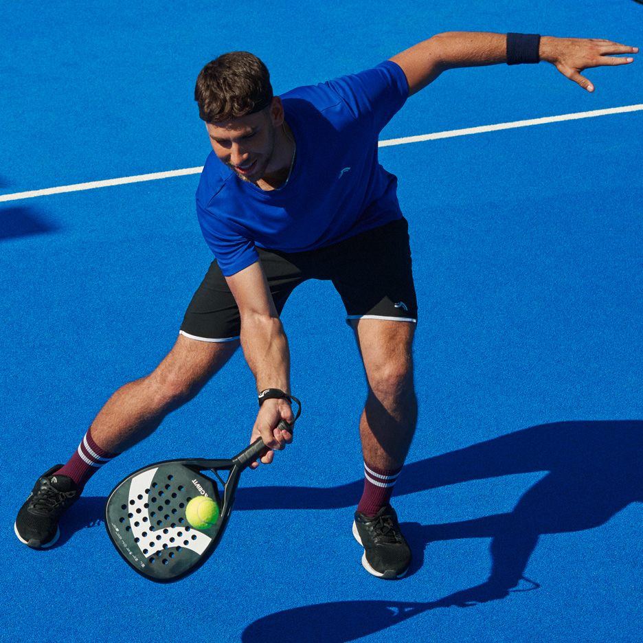 Muž v modrom tričku a čiernych šortkách hrá padel s raketou a loptičkou.